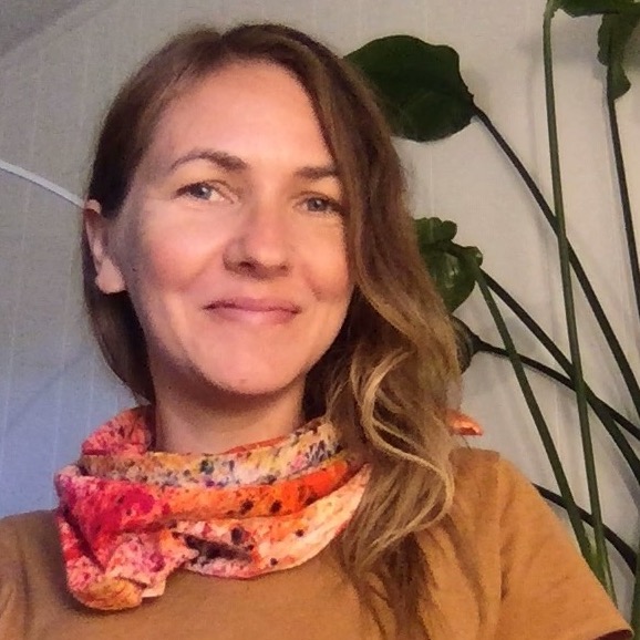 A headshot of a woman with long blonde hair wearing a brownish yellow tshirt and a red and pink floral silk scarf wrapped around her neck.