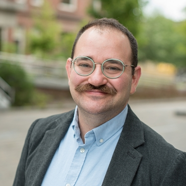 A headshot of a man with short dark hair, and a dark moustache. He is wearing a blue button up shirt and dark gray jacket